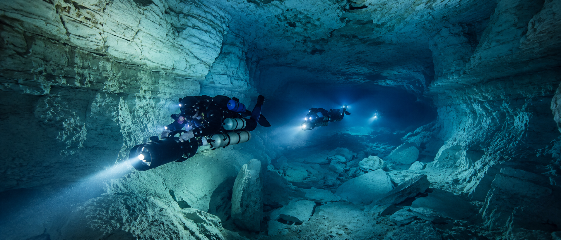 Technical cave divers riding DPV scooters through a vast illuminated underwater limestone cave tunnel